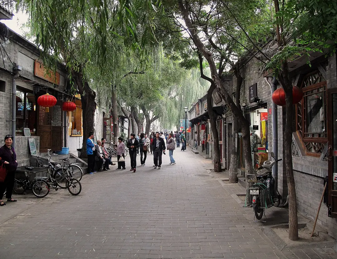 Tree-lined Nanluoguxiang lane with grey brick shopfronts, red lanterns and people walking