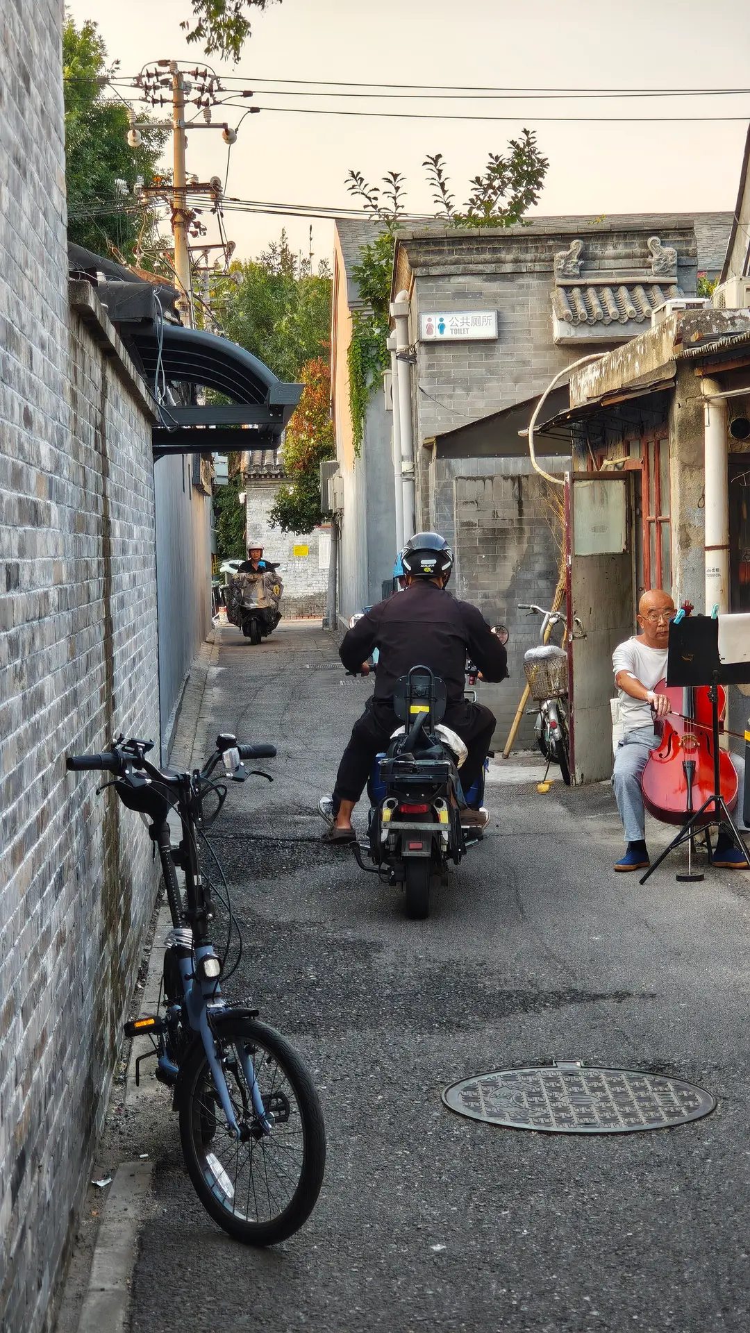 A narrow Beijing hutong with a scooter rider and a man playing the cello outside his door