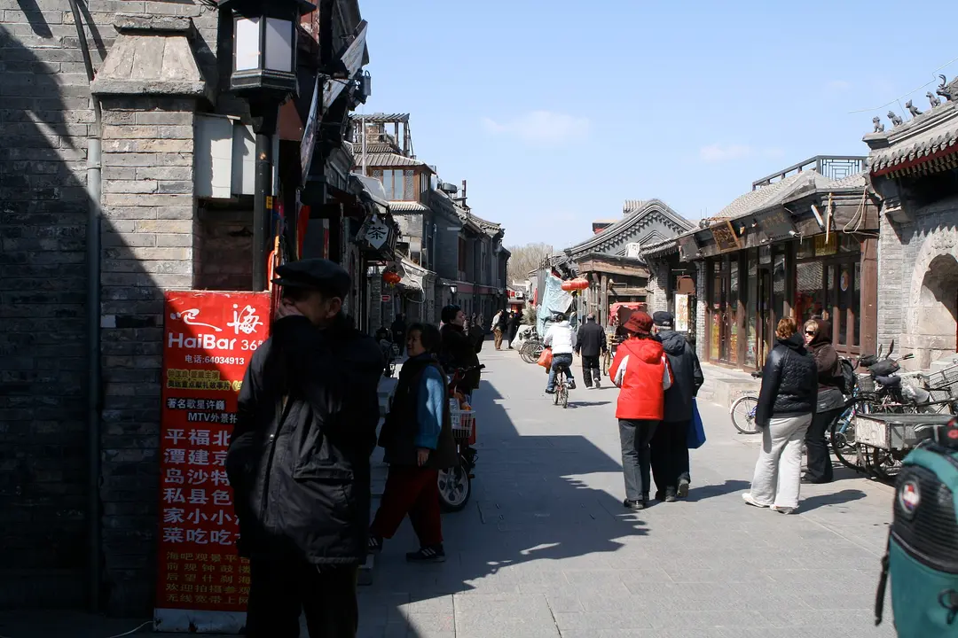 A grey brick hutong lane with shop signs and people walking in winter sunshine