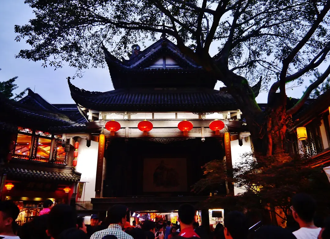 A lantern-lit wooden opera stage under a large tree on Jinli Street at dusk