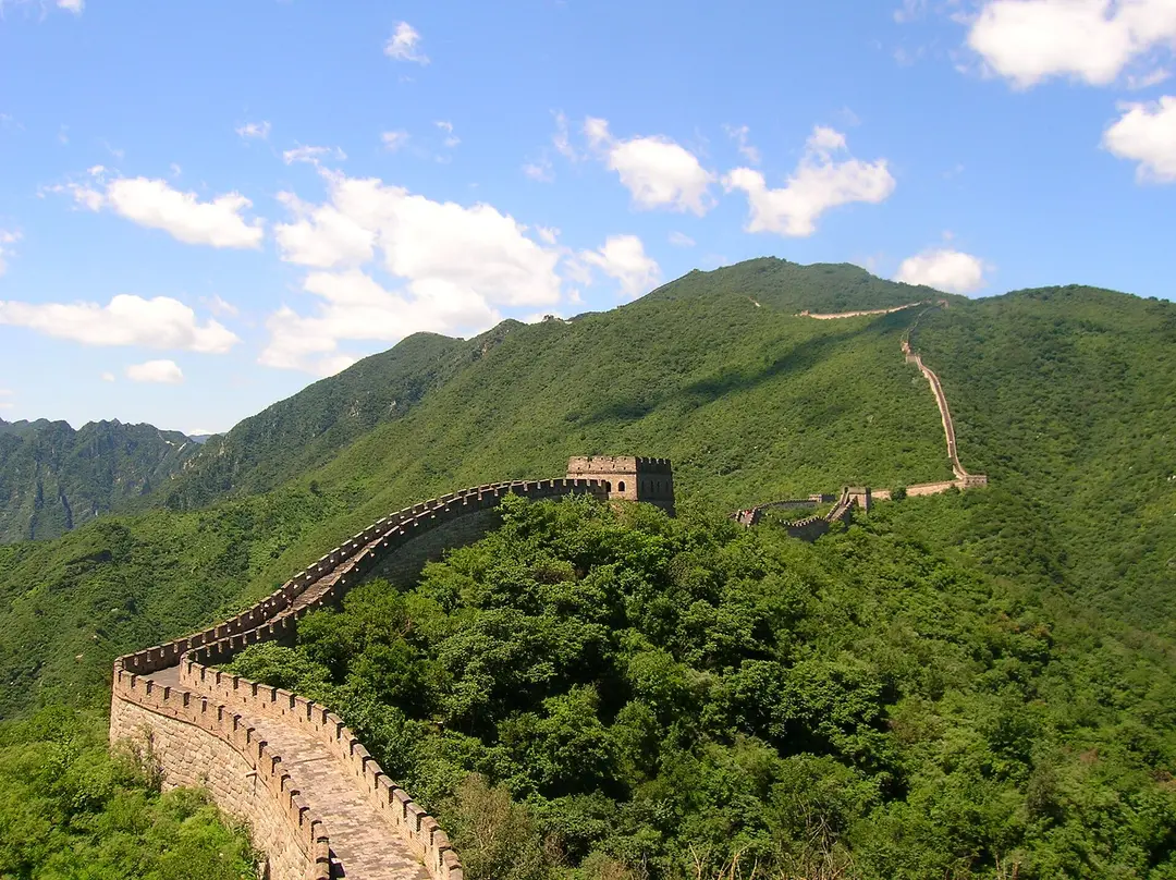 The Great Wall at Mutianyu running along a green, wooded ridge under a blue sky with white clouds