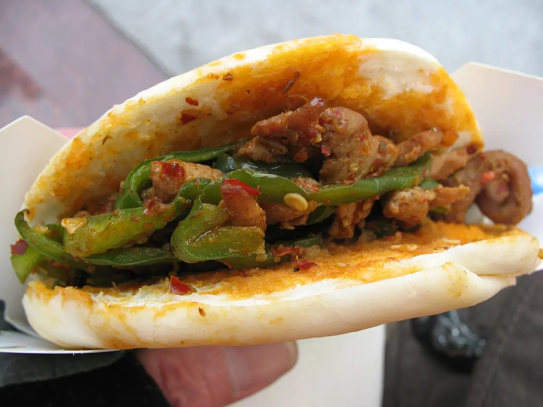 A roujiamo flatbread sandwich filled with chopped meat and green peppers, held in a paper wrapper