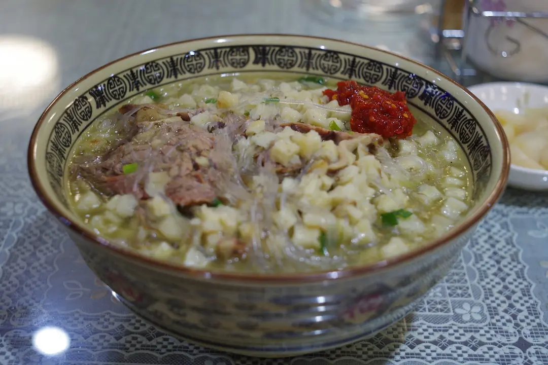 A bowl of lamb paomo: clear broth with sliced lamb, crumbled flatbread, glass noodles and chili paste