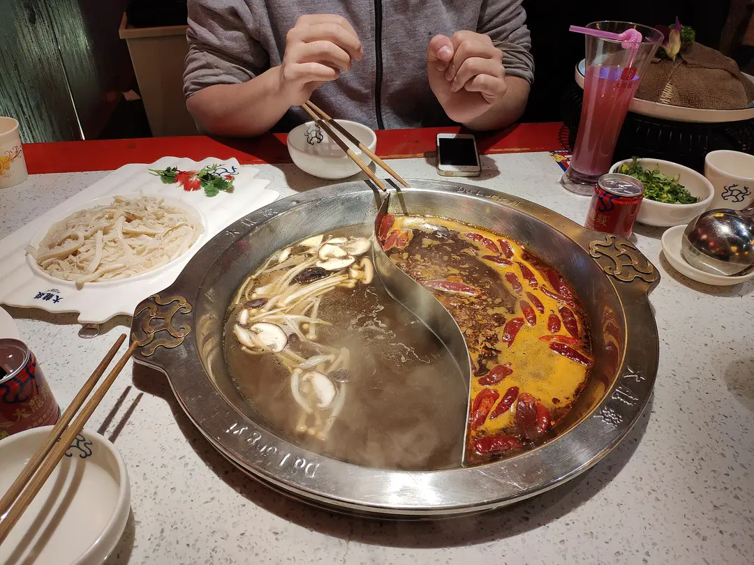 A split hotpot with red chili broth on one side and clear broth on the other, with noodles and side dishes