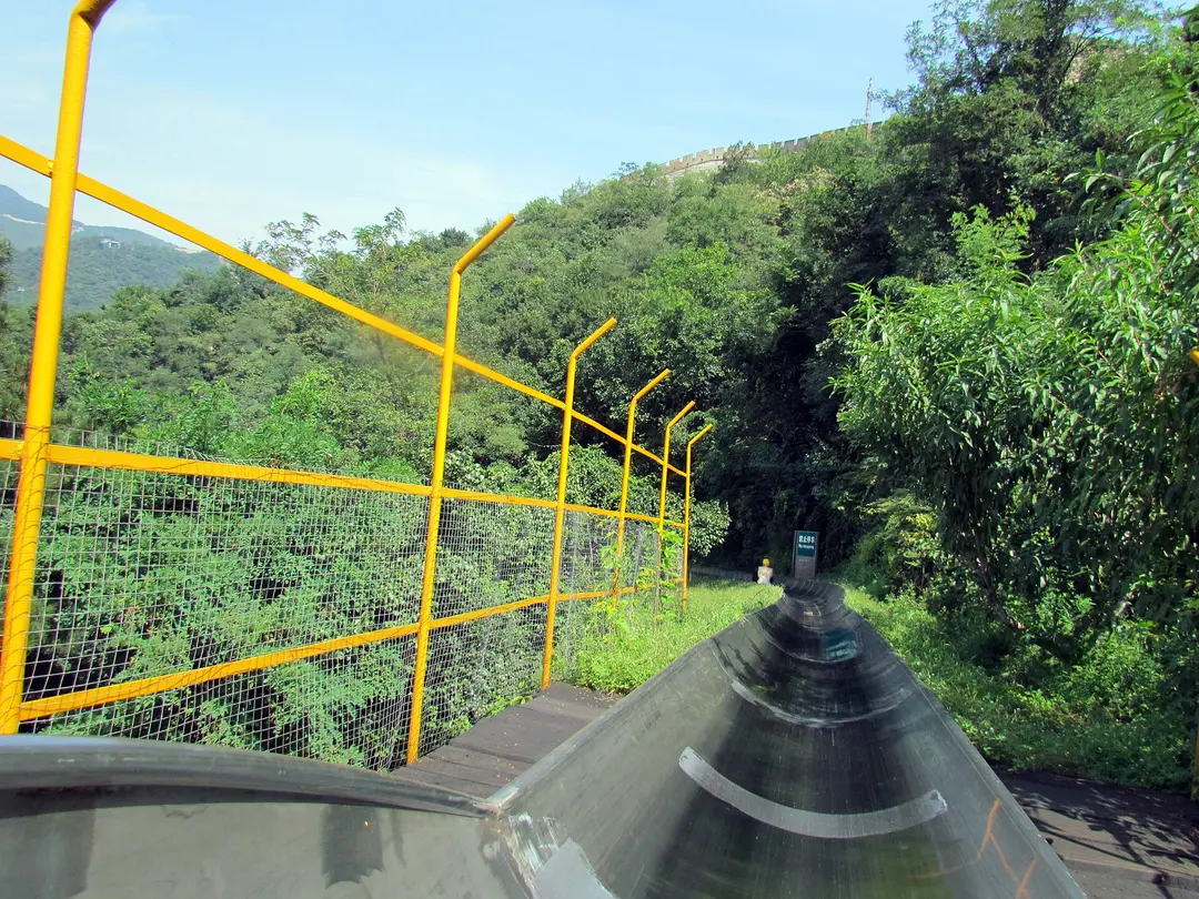 Metal toboggan track curving down through trees beside a yellow safety fence at Mutianyu